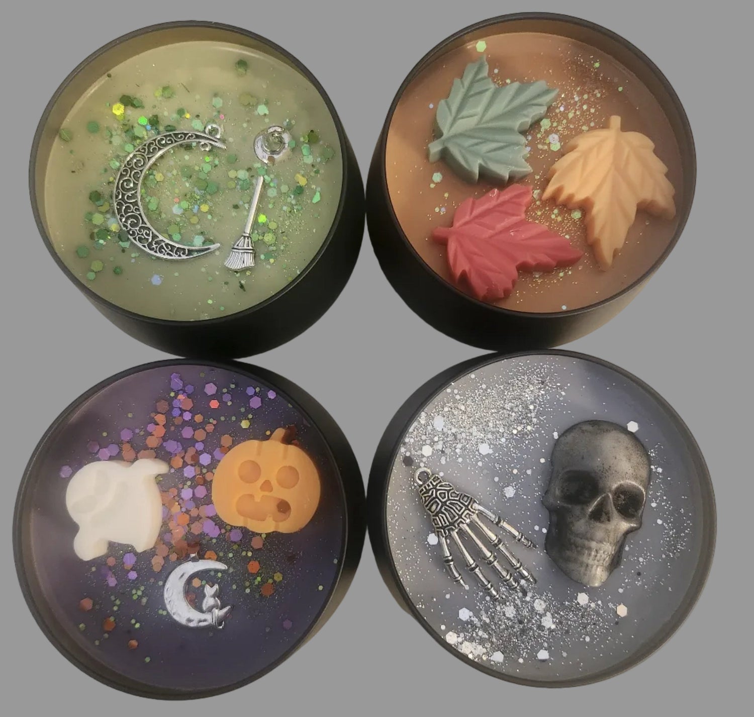 Four round decorative candles arranged in a 2x2 grid on a light beige textured background. Each candle has a dark rim and contains unique Halloween-themed or Fall-themed wax embeds.