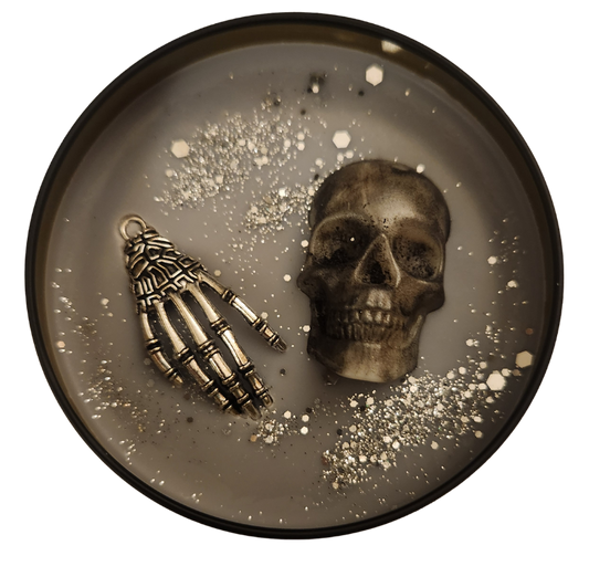 A black tin candle with grey wax and silver glitter with a wax skull head and a silver charm skull hand on a transparent background.