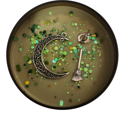 A black candle tin with a moss green wax and green shades of glitter with a silver charm half moon, broom, and witch hat on a transparent background.