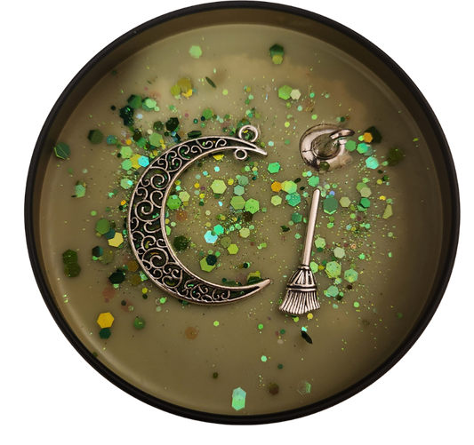 A black candle tin with a moss green wax and green shades of glitter with a silver charm half moon, broom, and witch hat on a transparent background.
