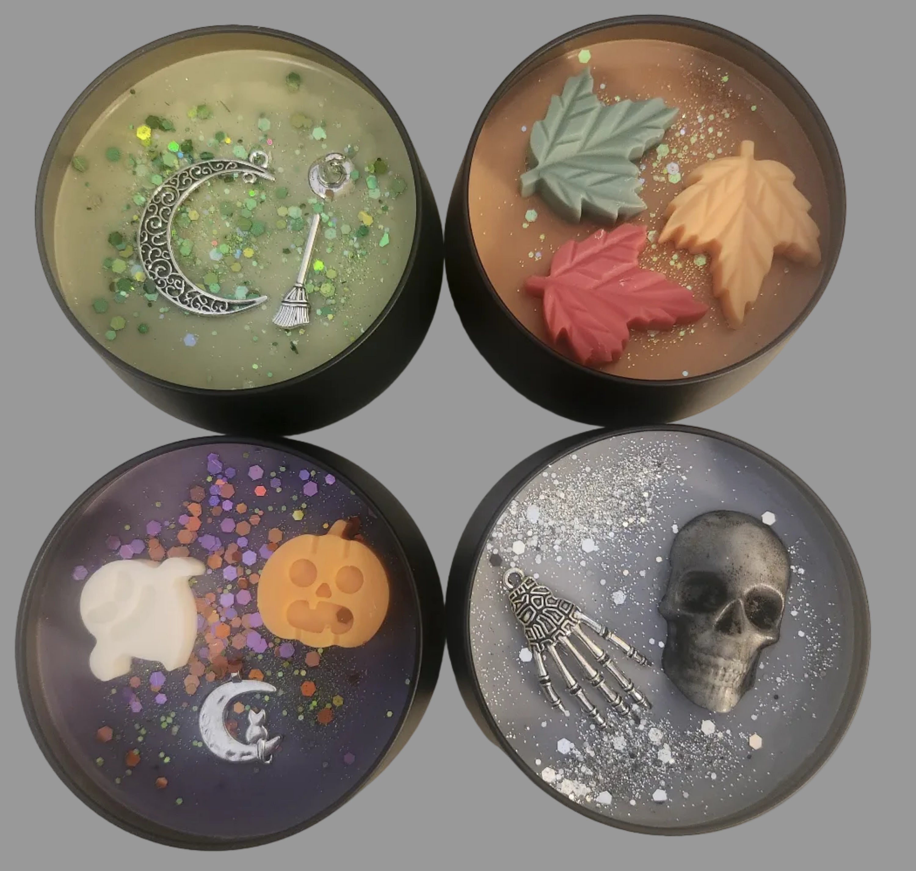 Four round decorative candles arranged in a 2x2 grid on a light beige textured background. Each candle has a dark rim and contains unique Halloween-themed resin designs: two fall leaves in orange and green on a warm brown candle, a skull and skeleton hand on a silver candle, a crescent moon and skull with green glitter on a yellow-green candle, and an orange skull, white ghost, and moon on a purple candle with multicolor glitter.