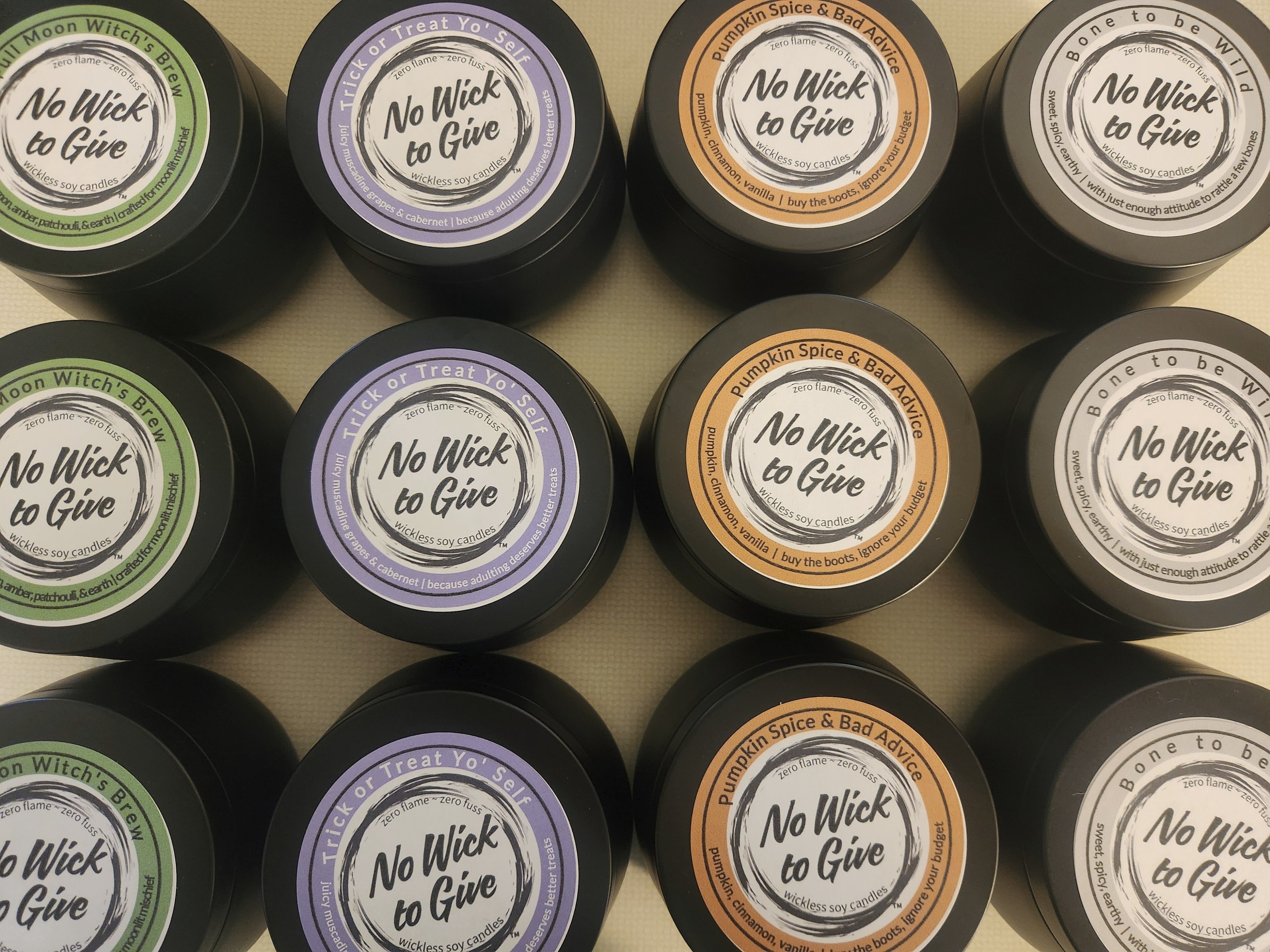 A grid of small black tins with round labels reading 'No Wick to Give' in different scent colors including green, lavender, orange, and white, arranged on a beige surface.