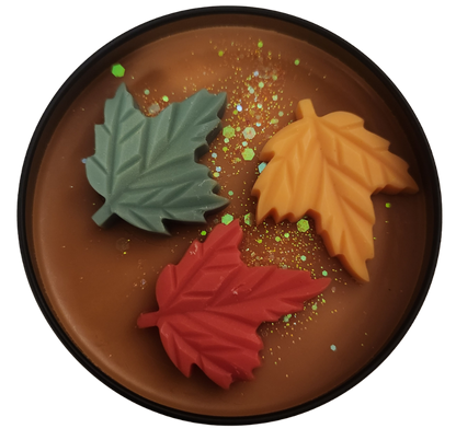 A black tin candle with pumpkin colored wax and a red, green, and orange leaves with golden glitter on top with a clear background.