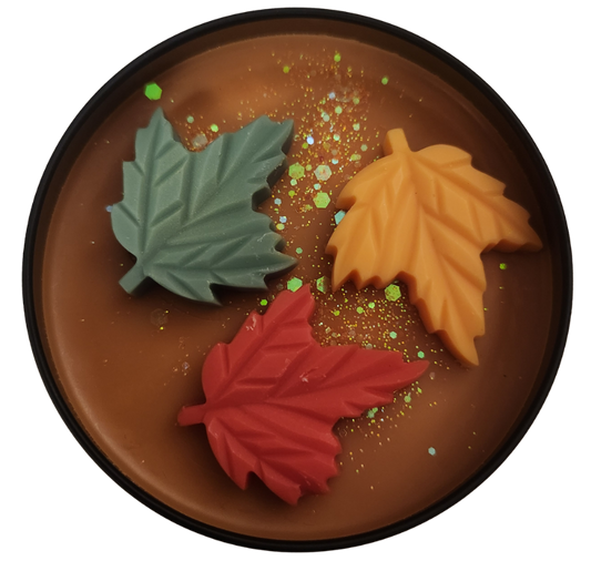 A black tin candle with pumpkin colored wax and a red, green, and orange leaves with golden glitter on top with a clear background.
