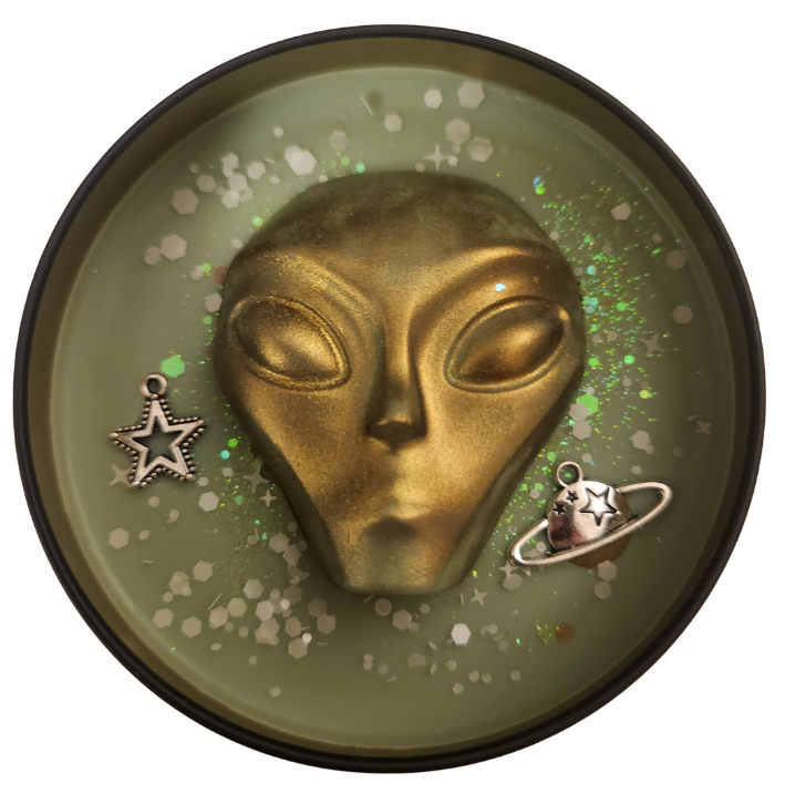 A black candle tin open to show a metallic gold alien head, scattered white and green glitter, and small celestial charms including a star and a ringed planet on green wax with a transparent background.