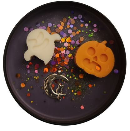 A black candle tin with purple wax featuring an off-white ghost-shaped mold, an orange pumpkin-shaped mold, and a silver crescent moon with a cat silhouette, surrounded by colorful confetti.