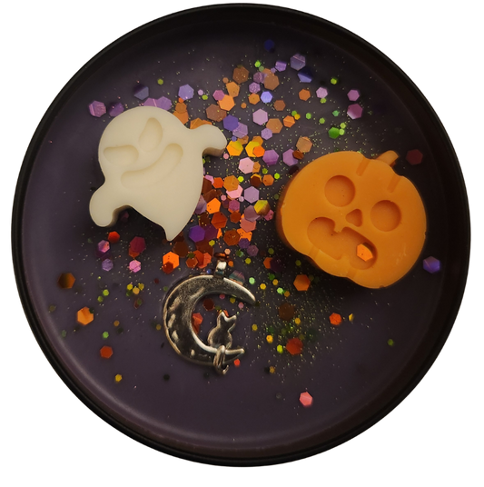 A black candle tin with purple wax featuring an off-white ghost-shaped mold, an orange pumpkin-shaped mold, and a silver crescent moon with a cat silhouette, surrounded by colorful confetti.